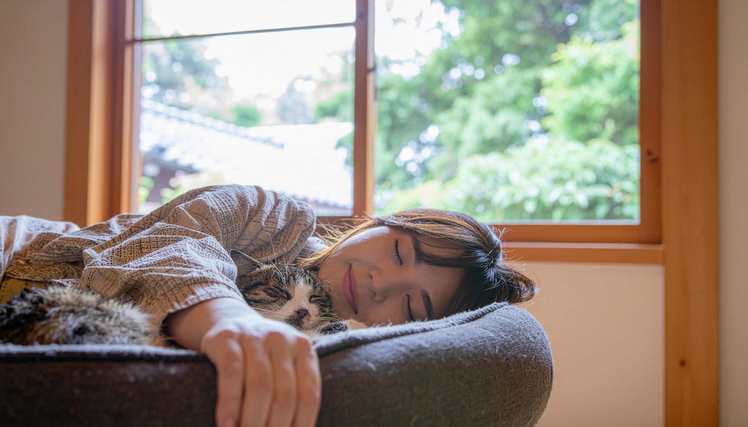 窓辺で愛猫と一緒に安心して眠る女性。猫と人が共に寝ることで深まる、スピリチュアルな絆と癒やしを象徴する光景。