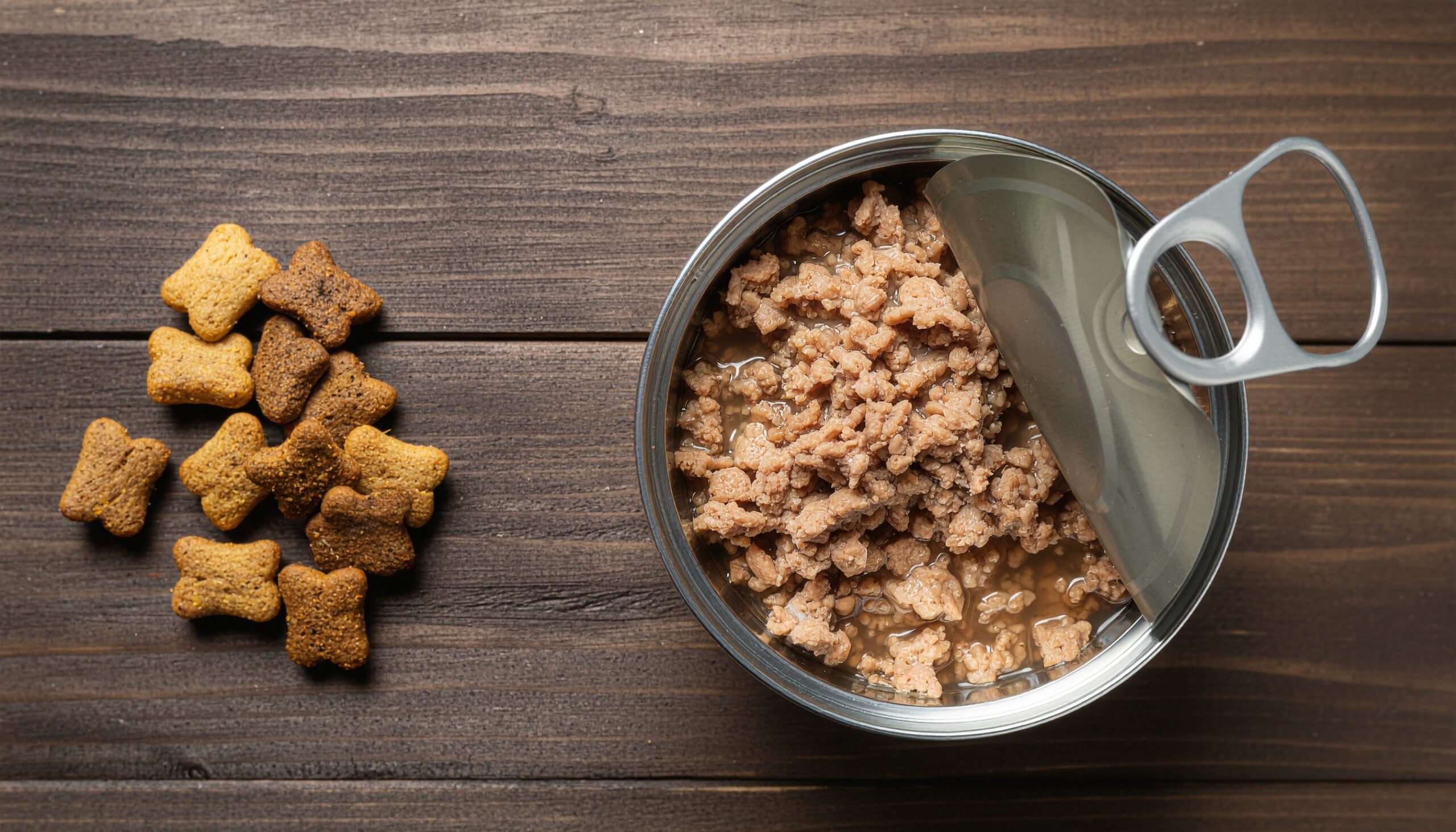 木目調のテーブルの上に、左側にカリカリのドライキャットフード、右側に開封されたウェットフードの缶詰が置かれており、それぞれのフードを真上から比較している写真。